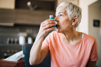 A person sits in a cushioned chair and holds a plastic breathing device to their mouth.