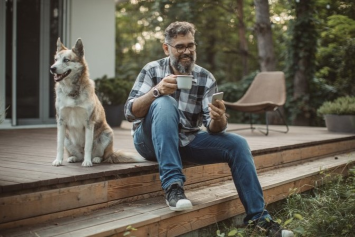 A person sits outside on a wooden deck with a Husky dog beside them and looks at their phone in one hand with a mug in the other. 