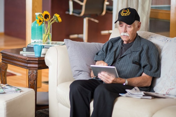 Older man wearing Vietnam Veteran hat sits on white couch with a tablet device in hand.